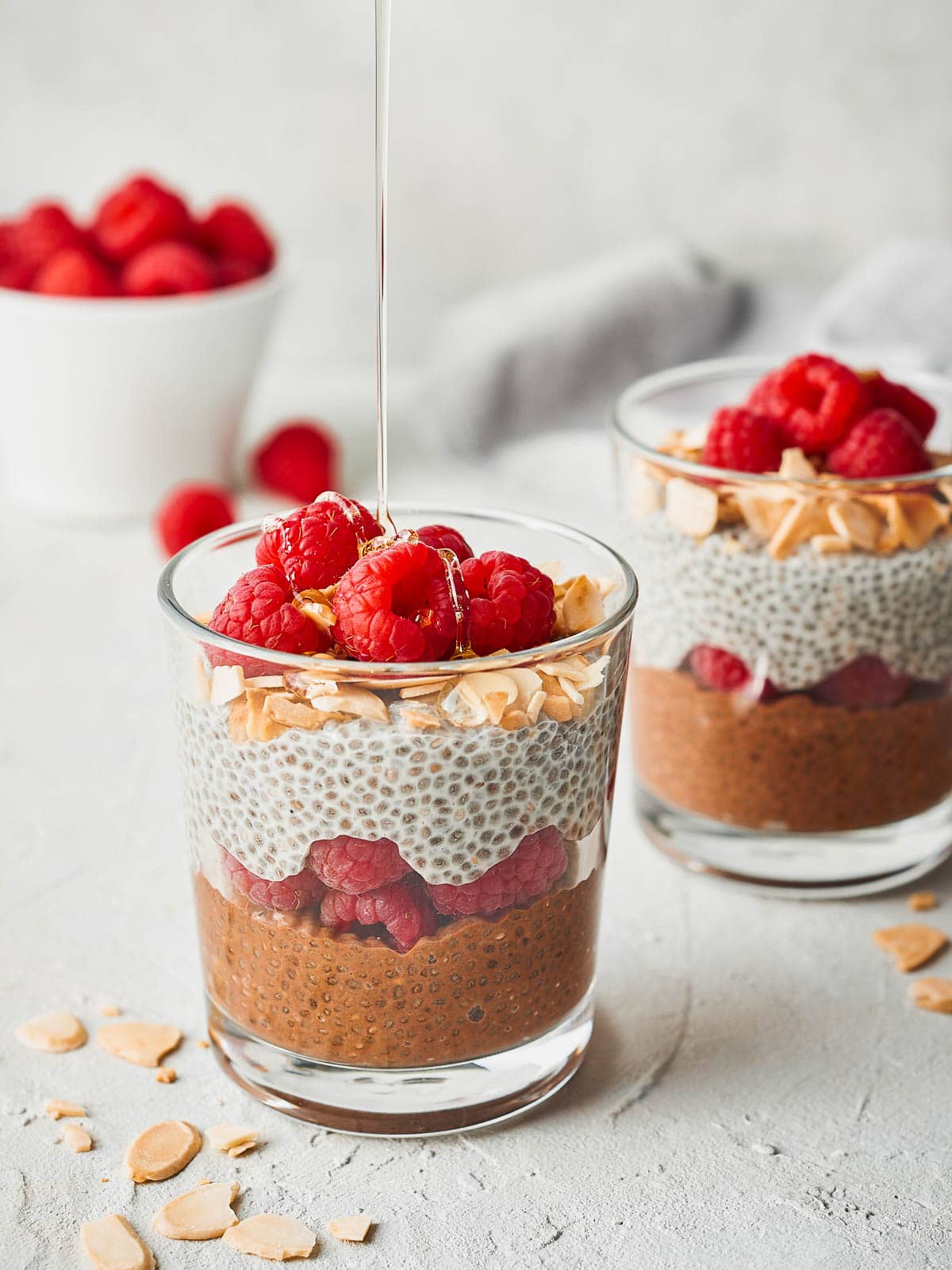 Two glasses of chia seed parfait, a small bowl of raspberries, with a drizzle of agave