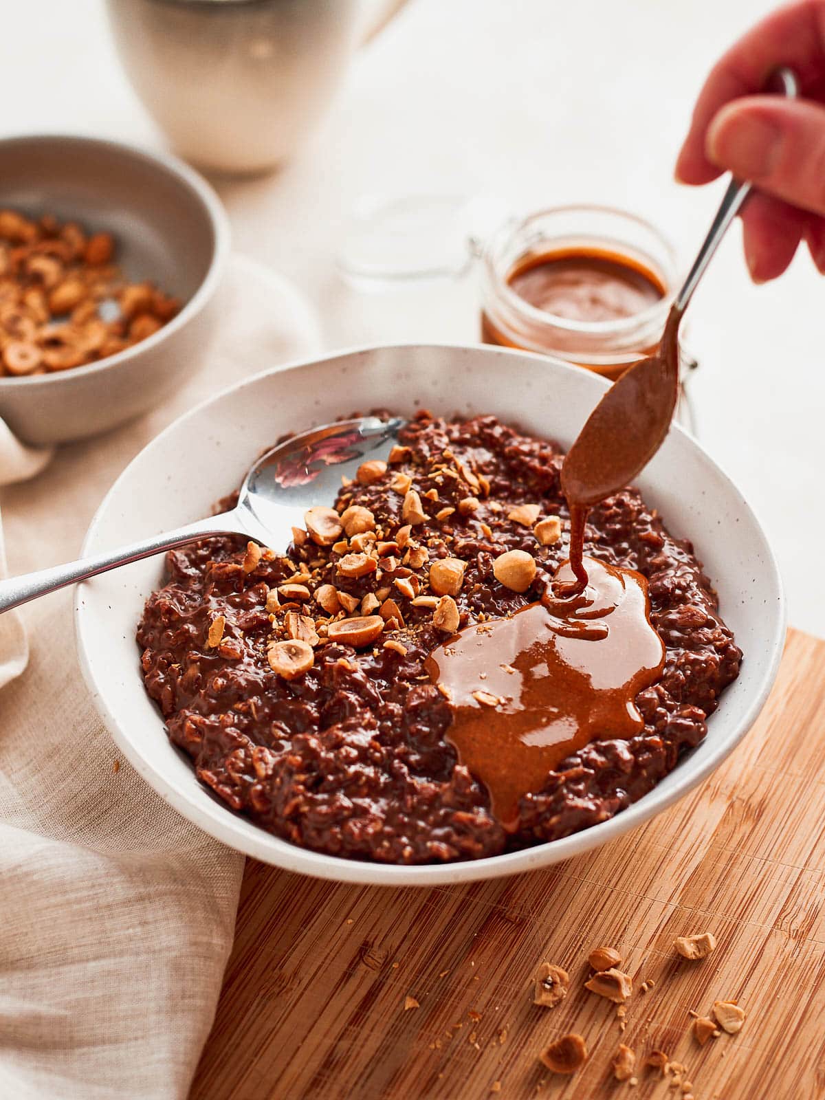 A bowl of chocolate hazelnut oatmeal with a mug and bowls of topping in the background, with hazelnut butter being drizzled on top