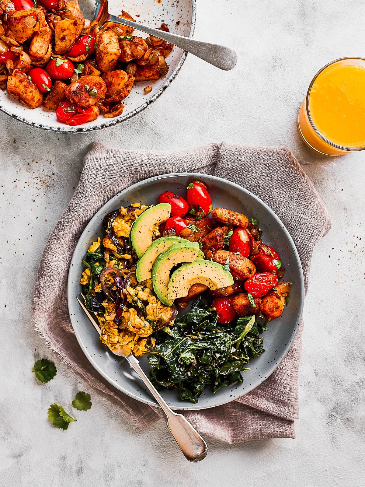 Mexican breakfast potatoes on a plate with sauteed kale and tofu scramble. A serving bowl of breakfast potatoes in the background.