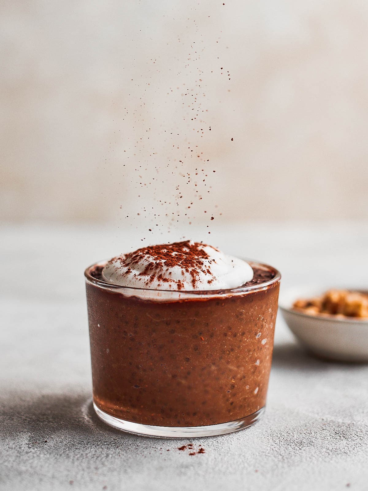 Some cocoa sprinkling down onto a glass of avocado chia pudding. A small bowl of nuts just behind.