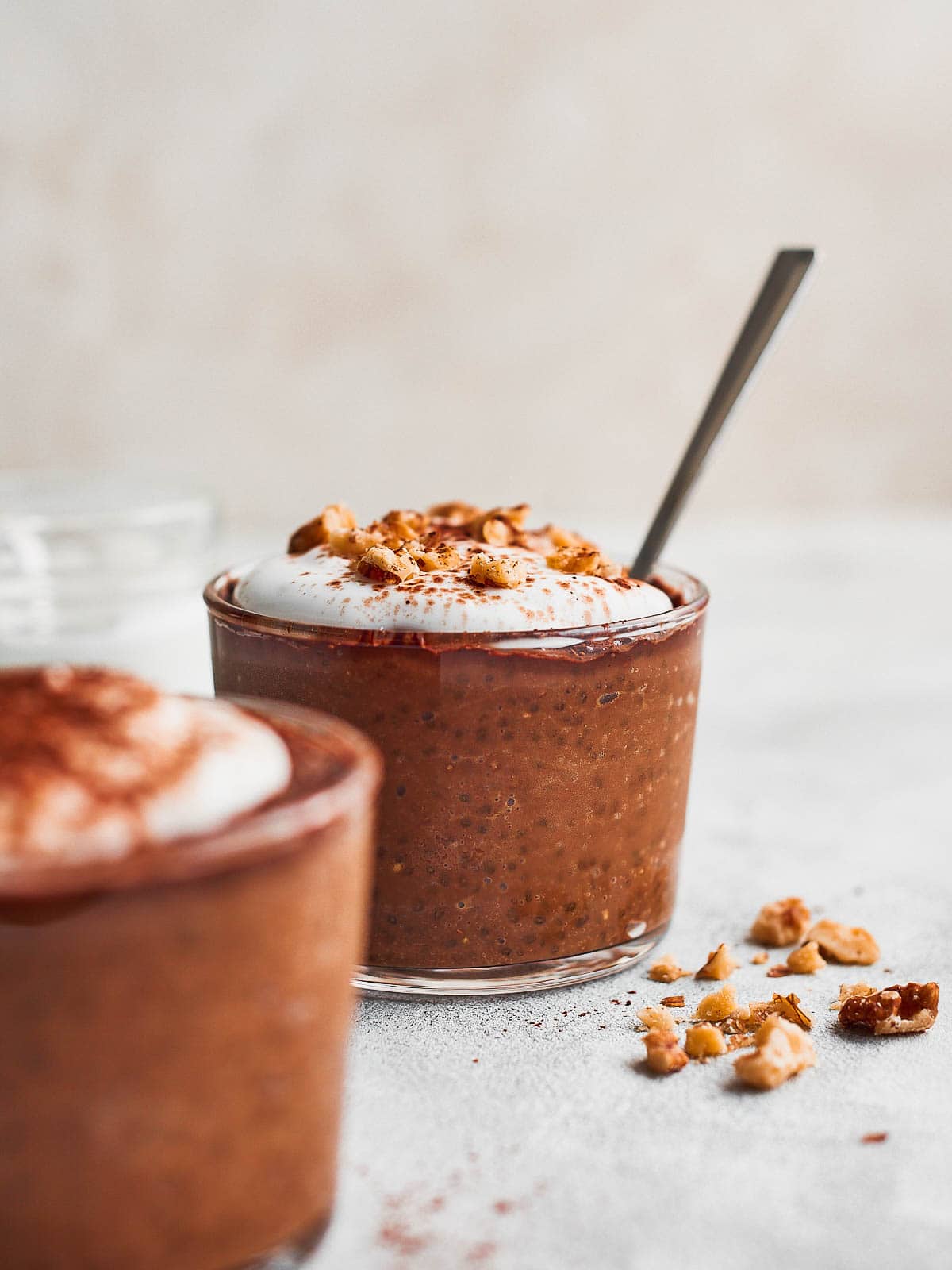 A glass of chocolate coffee chia pudding with a spoon coming out of it, with a pot of yoghurt in the background and another serving in the foreground